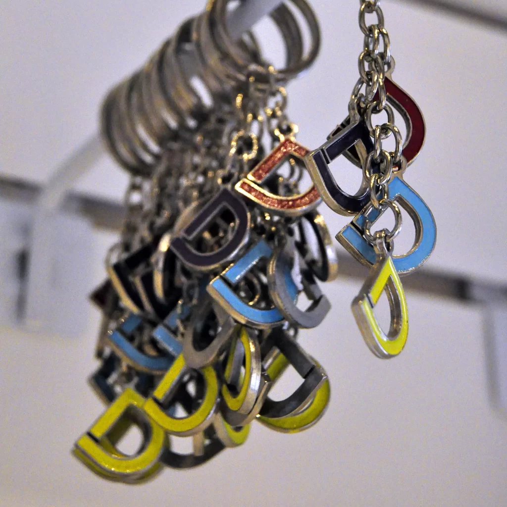 Primer plano de varios llaveros metálicos colgando, cada uno con un dije en forma de letra 'D' estilizada y rellena de esmalte de colores vivos como amarillo, azul y rojo.